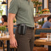 A man in a restaurant wearing a black leather belt holster with an Adyen POS handheld device, while holding a plate of food. The background shows a blurred dining area with tables and other diners.
