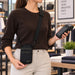 A woman wearing a brown ribbed sweater and beige pants models a black crossbody Adyen POS pouch with a black strap and subtle gold stitching in a retail store setting.