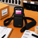 Black AgozTech Point Mobile crossbody holster with Point Mobile handheld device displayed on a warehouse packing desk, surrounded by a pick list clipboard, barcode labels, packing tape, and shipping box.