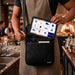 Restaurant server removing a PAX POS tablet from a black Agoz crossbody holster with adjustable shoulder sling and front storage pocket.