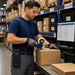 Warehouse worker sealing a cardboard box with a tape gun while wearing a compact black AGOZ belt case holding a Honeywell Dolphin CK65 at a packing station.