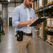 A man in a warehouse wearing a light blue button-down shirt, khaki pants, and a black utility belt pouch with a small orange accent, holding a clipboard and writing. He wears a Wasp scanner handle belt holster. 